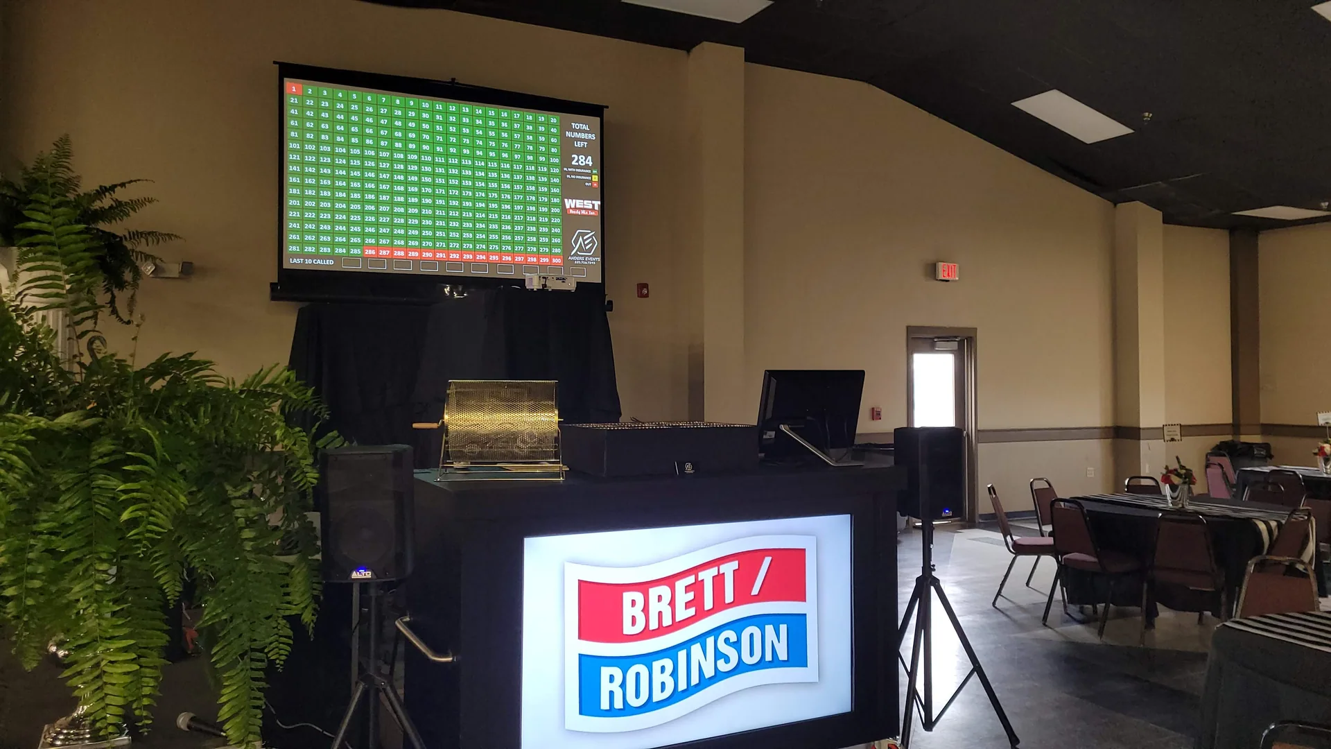 A sponsor logo for Brett/Robinson displayed on the screen built into the booth front, with the number drawing board projected above and a brass ticket hopper alongside.