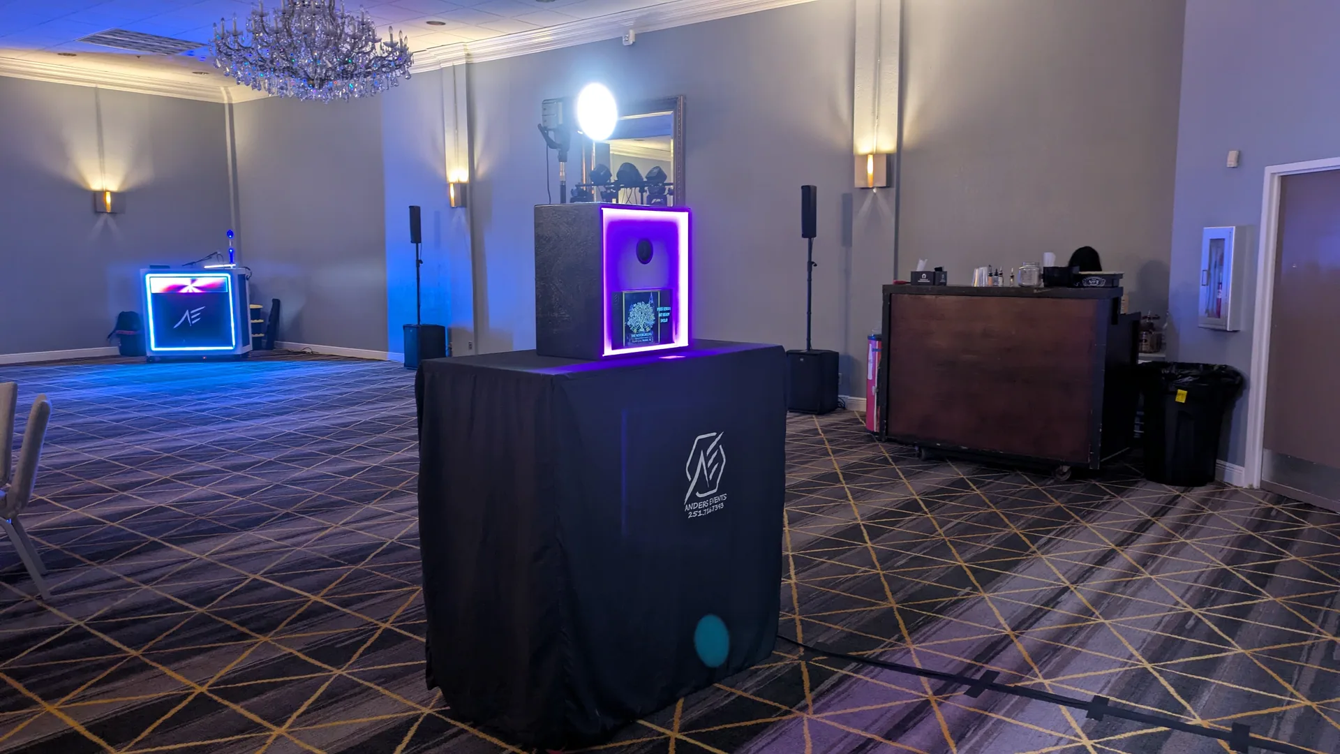 The photo booth in a hotel ballroom under a chandelier, its magenta LED surround lit, standing on a branded Anders Events cover with the DJ booth glowing cyan behind it.