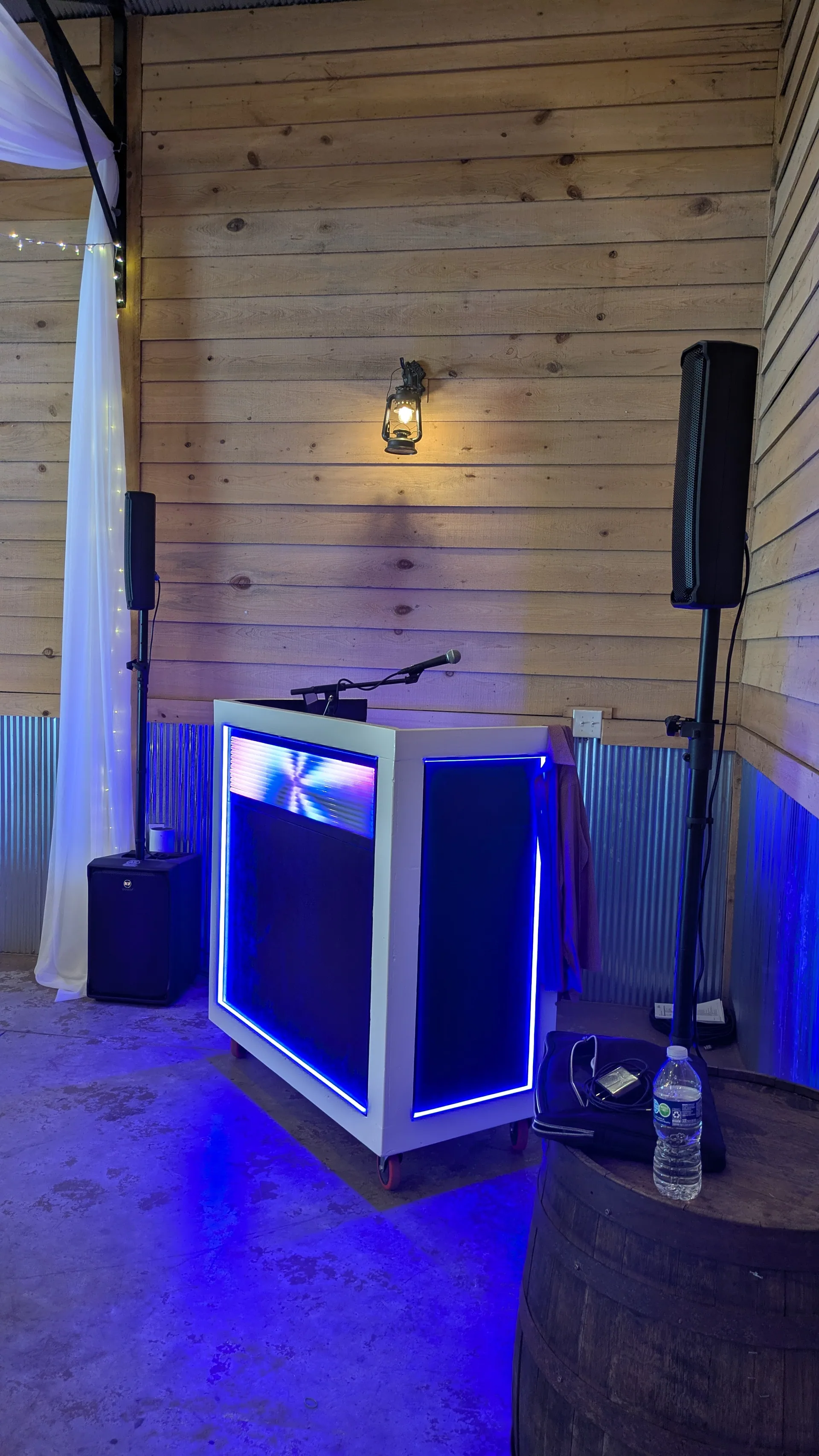A custom Anders Events DJ booth glowing with a blue LED perimeter inside a wooden barn venue, flanked by PA speakers on stands.