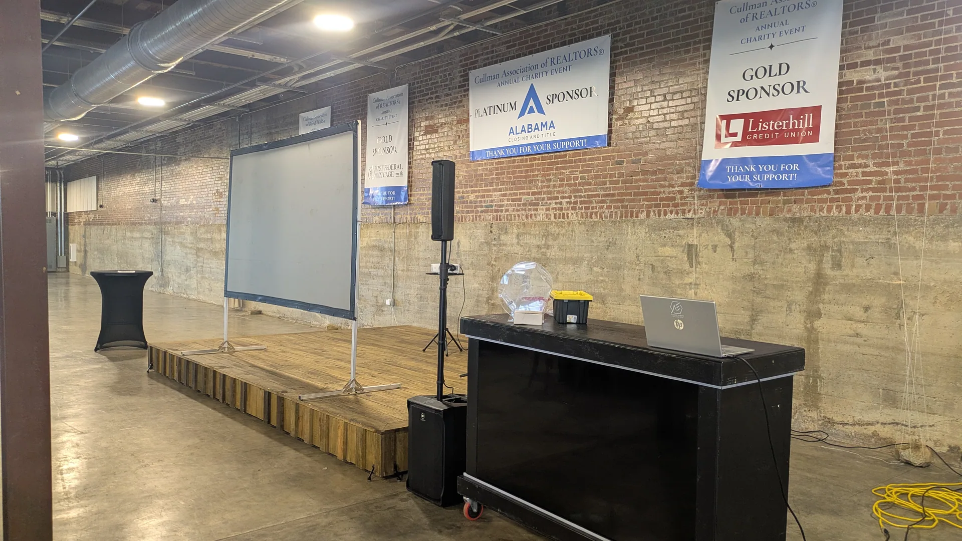 A charity fundraiser set up in a brick venue: projection screen, PA, ticket hopper and booth in position before guests arrive.